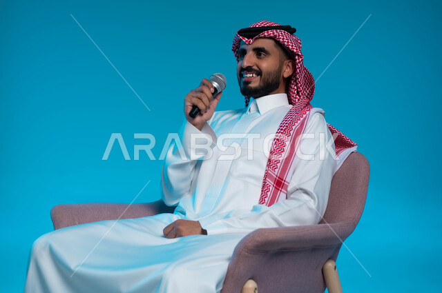 Enjoying the performance of vocal talents, a portrait of a Saudi Gulf Arab young man wearing traditional clothing, holding a microphone in his hand and singing enthusiastically with gestures indicating integration and interaction, practicing the talent of singing, recording a singing audio clip, blue background
