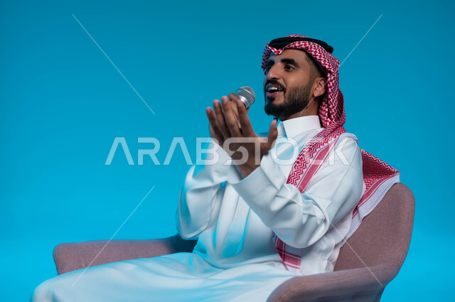 ممارسة موهبة الغناء، الاستمتاع بأداء المواهب الصوتية، بورتريه لشاب عربي خليجي سعودي يرتدي الزي التقليدي يمسك بيده المايكروفون ويغني بحماس بإيماءات تدل على الاندماج والتفاعل، تسجيل مقطع صوتي غنائي، خلفية زرقاء