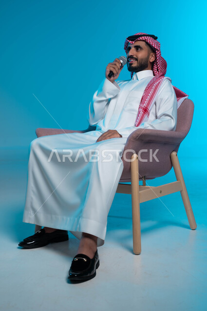 الاستمتاع بأداء المواهب الصوتية، بورتريه لشاب عربي خليجي سعودي يرتدي الزي التقليدي يمسك بيده المايكروفون ويغني بحماس بإيماءات تدل على الاندماج والتفاعل، ممارسة موهبة الغناء، تسجيل مقطع صوتي غنائي، خلفية زرقاء