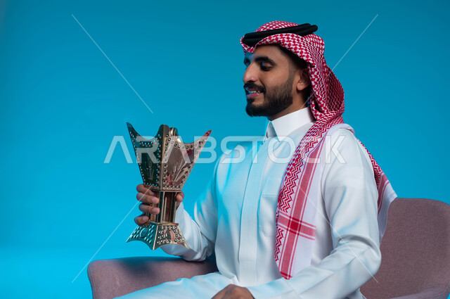 The use of agarwood for holidays and occasions, Saudi customs and traditions, perfuming with agarwood and incense, a close-up portrait of a smiling Saudi Arabian Gulf man wearing a traditional thobe and shemagh, perfumed from the incense burner holder, taking care of the external appearance, blue background