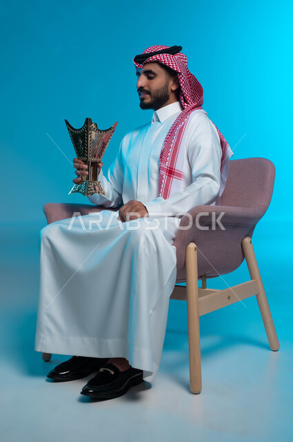 الاستمتاع برائحة العطور والبخور, العادات والتقاليد السعودية, استخدام خشب العود بالأعياد والمناسبات, بورتريه لشاب عربي خليجي سعودي يرتدي الشماغ والثوب التقليدي يحمل المبخرة, الأناقة والاهتمام بالمظهر الخارجي, خلفية زرقاء