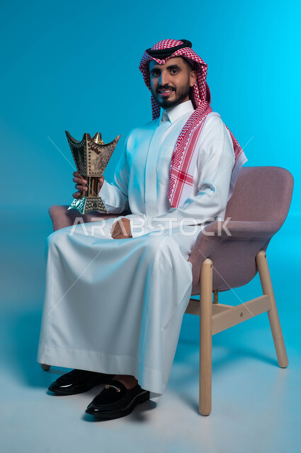 استخدام خشب العود بالأعياد والمناسبات, العادات والتقاليد السعودية, التطيب والتعطر بالبخور, بورتريه لرجل عربي خليجي سعودي يرتدي الثوب التقليدي والشماغ يحمل بيده حامل المبخرة وينظر إلى الكاميرا, الاهتمام بالمظهر الخارجي, خلفية زرقاء