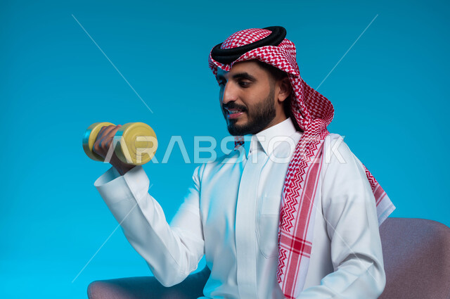 نشاط بدني للمحافظة على الصحة واللياقة الجسدية، اتباع أسلوب حياة صحي، بورتريه مقرب لشاب عربي خليجي سعودي يرتدي الزي التقليدي يمارس التمارين الرياضية مع الدمبلز، كمال الأجسام وتقوية وبناء العضلات، رفع الأثقال والأوزان الحديدية الثقيلة، خلفية زرقاء.