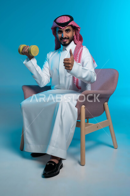 Strengthening and building muscles, physical activity to maintain health and physical fitness, following a healthy lifestyle, close-up portrait of a Saudi Gulf Arab young man wearing traditional clothing exercising with dumbbells, pointing with gestures of quality and admiration, lifting heavy iron weights, blue background.