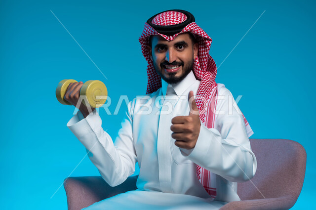 Strengthening and building muscles, physical activity to maintain health and physical fitness, following a healthy lifestyle, close-up portrait of a Saudi Gulf Arab young man wearing traditional clothing exercising with dumbbells, pointing with gestures of quality and admiration, lifting heavy iron weights, blue background.