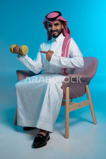 ممارسة التمارين الرياضية لتقوية وبناء العضلات، نشاط بدني للمحافظة على الصحة واللياقة الجسدية، اتباع أسلوب حياة صحي، بورتريه لشاب عربي خليجي سعودي يرتدي الزي التقليدي يشير بإصبع السبابة إلى الدمبلز، رفع الأوزان الحديدية الثقيلة، خلفية زرقاء.
