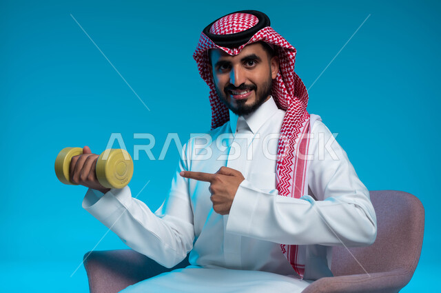 ممارسة التمارين الرياضية لتقوية وبناء العضلات، نشاط بدني للمحافظة على الصحة واللياقة الجسدية، اتباع أسلوب حياة صحي، بورتريه لشاب عربي خليجي سعودي يرتدي الزي التقليدي يشير بإصبع السبابة إلى الدمبلز، رفع الأوزان الحديدية الثقيلة، خلفية زرقاء.