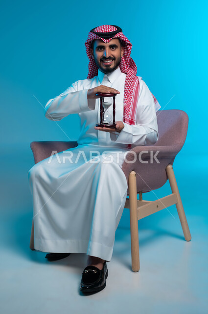 القيام بمراقبة توقيت الرمل, مفهوم إدارة الأعمال وتنظيم الجدول الزمني, إدراك قيمة اللحظات الثمينة, بورتريه لشاب عربي خليجي سعودي مبتسم يرتدي الزي التقليدي يمسك بيده الساعة الرملية الزجاجية (البنكام), النظر إلى الكاميرا بإيماءات السرور, خلفية زرقاء