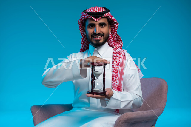 القيام بمراقبة توقيت الرمل, مفهوم إدارة الأعمال وتنظيم الجدول الزمني, إدراك قيمة اللحظات الثمينة, بورتريه لشاب عربي خليجي سعودي مبتسم يرتدي الزي التقليدي يمسك بيده الساعة الرملية الزجاجية (البنكام), النظر إلى الكاميرا بإيماءات السرور, خلفية زرقاء