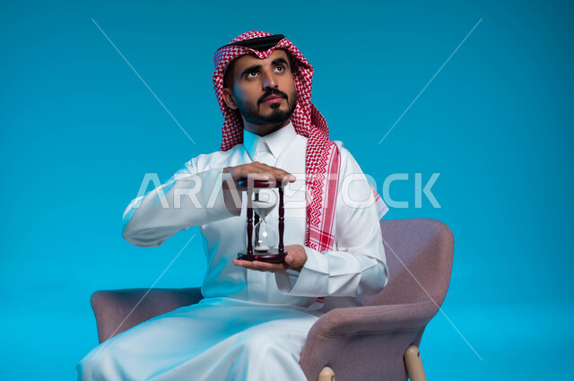 إدراك قيمة اللحظات الثمينة, القيام بمراقبة توقيت الرمل, مفهوم إدارة الأعمال وتنظيم الجدول الزمني, بورتريه لشاب عربي خليجي سعودي يرتدي الزي التقليدي يمسك بيده الساعة الرملية الزجاجية (البنكام) وينظر إلى الأعلى, خلفية زرقاء