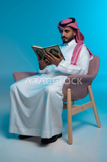 Reciting the Book of God Almighty, diligence and humility in worship, drawing closer to the Lord of the Worlds by performing obligations and acts of worship, portrait of a Saudi Gulf Arab young man wearing a shemagh and traditional dress, reading the Holy Quran, blue background