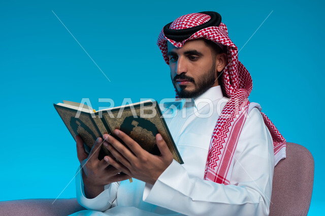 التقرب إلى رب العالمين بأداء الفرائض والعبادات, تلاوة كتاب الله تعالى ,الاجتهاد و الخشوع في العباده, بورتريه لشاب عربي خليجي سعودي يرتدي الشماغ والثوب التقليدي يقوم يقراءة القرآن الكريم, خلفية زرقاء