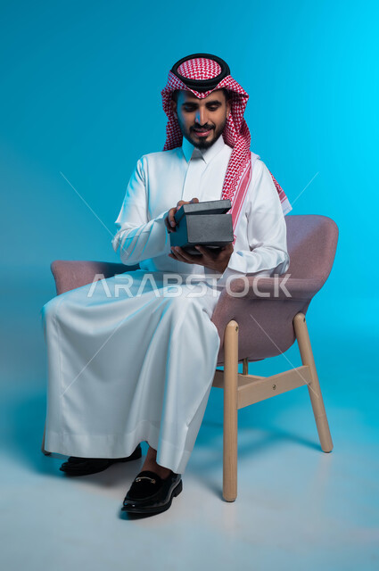 تقديم الهدايا في الأعياد والمناسبات, الجلوس بإيماءات السرور والفرح, بورتريه لشاب عربي خليجي سعودي مبتسم يرتدي الشماغ والثوب التقليدي يقوم بفتح علبة الهدية بإيماءات السعادة, خلفية زرقاء