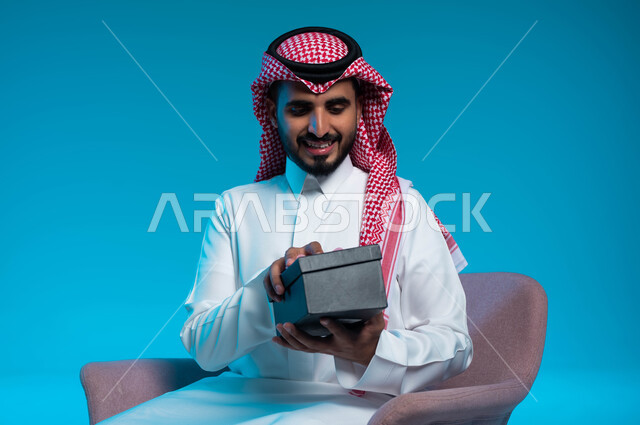 الجلوس بإيماءات السرور والفرح, تقديم الهدايا في الأعياد والمناسبات, بورتريه لشاب عربي خليجي سعودي مبتسم يرتدي الشماغ والثوب التقليدي يقوم بفتح علبة الهدية بإيماءات السعادة, خلفية زرقاء
