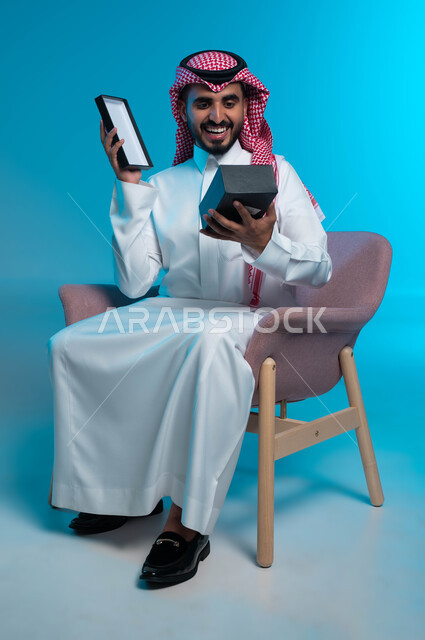 الفرح والسعادة بالحصول على الهدية, الجلوس بإيماءات السرور والاندهاش, تقديم الهدايا في الأعياد والمناسبات, بورتريه لشاب عربي خليجي سعودي مبتسم يرتدي الشماغ والثوب التقليدي يمسك بيده علبة, خلفية زرقاء