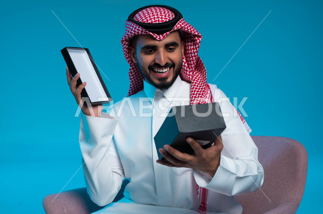 الجلوس بإيماءات السرور والاندهاش, الفرح والسعادة بالحصول على الهدية, تقديم الهدايا في الأعياد والمناسبات, بورتريه مقرب لشاب عربي خليجي سعودي مبتسم يرتدي الشماغ والثوب التقليدي يمسك بيده علبة, خلفية زرقاء.