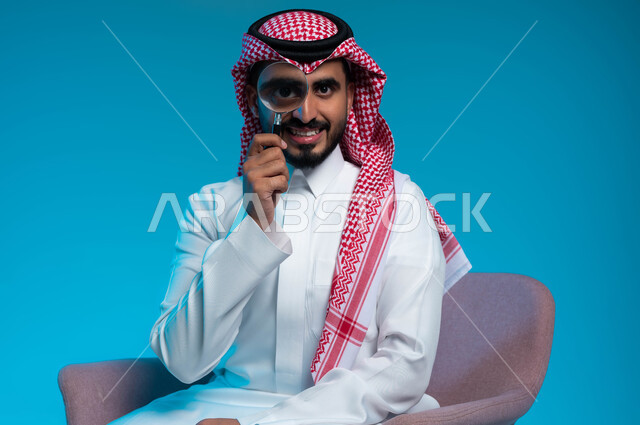 مفهوم الفحص والاستكشاف والقراءة عن قرب، أدوات بصرية للتدقيق والتركيز، بورتريه مقرب لرجل عربي خليجي سعودي مبتسم يرتدي الزي التقليدي يحمل بيده عدسة مكبرة لتوضيح وتحسين رؤية شيء ما، خلفية زرقاء