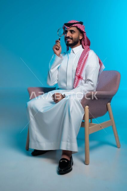 أدوات بصرية للتدقيق والتركيز، بورتريه لرجل عربي خليجي سعودي مبتسم يرتدي الزي التقليدي يحمل بيده عدسة مكبرة لتوضيح وتحسين رؤية شيء ما، مفهوم الفحص والاستكشاف والقراءة عن قرب، خلفية زرقاء