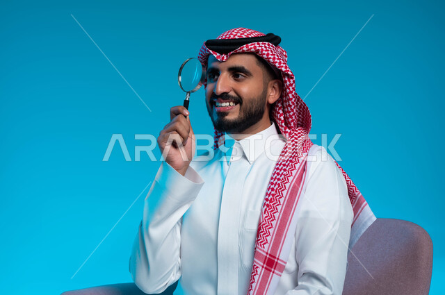 مفهوم الفحص والاستكشاف والقراءة عن قرب، أدوات بصرية للتدقيق والتركيز، بورتريه مقرب لرجل عربي خليجي سعودي مبتسم يرتدي الزي التقليدي يحمل بيده عدسة مكبرة لتوضيح وتحسين رؤية شيء ما، خلفية زرقاء