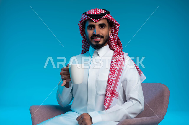 الحيوية والنشاط عند شرب الكافيين، الشعور والاحساس بالدفء عند تناول المشروبات الساخنة، بورتريه لرجل عربي سعودي خليجي يرتدي الزي التقليدي يحمل بيده كوب القهوة وينظر إلى الكاميرا، خلفية زرقاء
