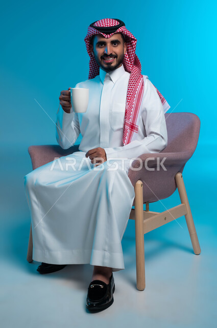 النظر إلى الكاميرا بإيماءات الفرح والاستمتاع، الشعور والاحساس بالدفء عند تناول المشروبات الساخنة، الحيوية والنشاط عند شرب الكافيين، بورتريه لرجل عربي سعودي خليجي مبتسم يرتدي الزي التقليدي يحمل بيده كوب القهوة، خلفية زرقاء