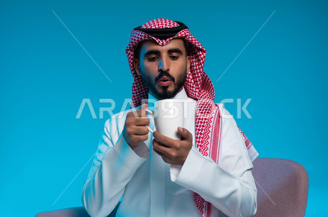قضاء الوقت بشرب المشروب المفضل، تعديل المزاج بتناول الكافيين، احتساء المشروبات الساخنة والأعشاب الشتوية الدافئة, بورتريه مقرب لرجل عربي خليجي سعودي يرتدي الثوب والشماغ التقليدي يحاول التخفيف من سخونة القهوة, خلفية زرقاء