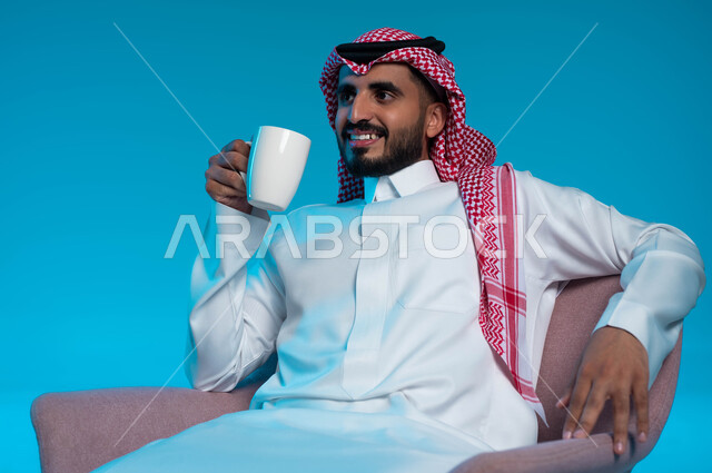 الجلوس والاستمتاع بالمشروبات الساخنة والأعشاب الشتوية الدافئة, قضاء وقت الفراغ باحتساء المشروب المفضل, بورتريه لرجل عربي خليجي سعودي مبتسم يرتدي الثوب والشماغ التقليدي يقوم بشرب القهوة ومشاهدة شيء ما بإيماءات السعادة, خلفية زرقاء