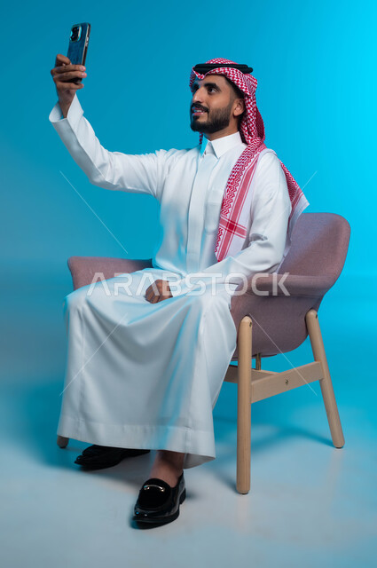 Documenting happy moments, using modern devices and technologies, a portrait of a smiling Saudi Arabian Gulf man wearing a shemagh and traditional thobe, taking selfies on a mobile phone with happy gestures, blue background