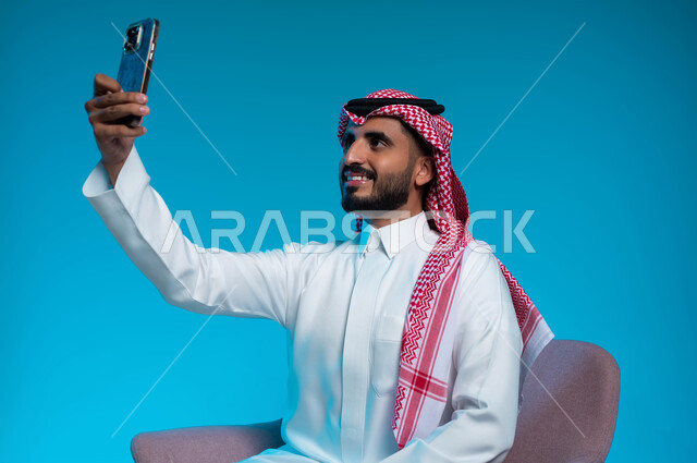 توثيق اللحظات السعيدة والمميزة, استخدام الاجهزة والتقنيات الحديثة, بورتريه مقرب لرجل عربي سعودي خليجي مبتسم يرتدي الشماغ والثوب التقليدي يقوم بالتقاط صور السيلفي عبر الهاتف المحمول بإيماءات السرور, خلفية زرقاء