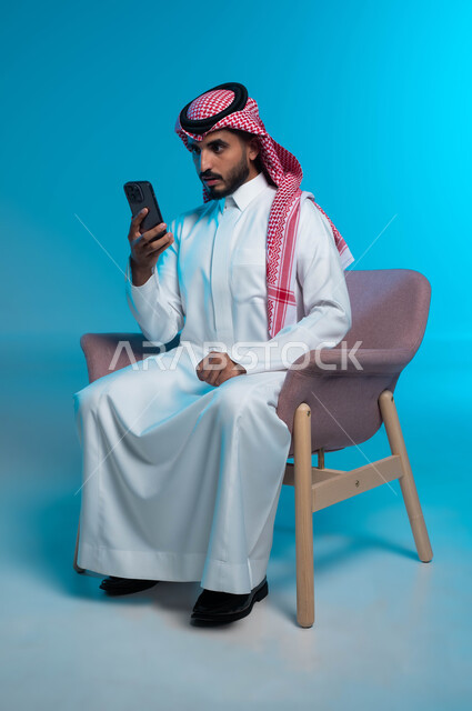 تعابير وإيماءات الصدمة والمفاجأة لرؤية شيء ما عبر الجوال, بورتريه لشاب عربي سعودي خليجي يرتدي الشماغ والثوب التقليدي يستخدم الهاتف المحمول، استخدام الأجهزة والتقنيات الحديثة والمتطورة, خلفية زرقاء