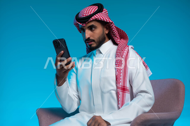 إيماءات وتعابير الصدمة والمفاجأة لرؤية شيء ما عبر الجوال, بورتريه مقرب لشاب عربي سعودي خليجي يرتدي الشماغ والثوب التقليدي يستخدم الهاتف المحمول، استخدام الأجهزة والتقنيات الحديثة والمتطورة, خلفية زرقاء