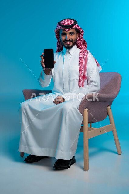 Gestures of joy and happiness using modern and advanced devices and technologies, a portrait of a smiling Saudi Gulf Arab man wearing a traditional thobe and shemagh, displaying a mobile phone with a blank black screen, blue background.