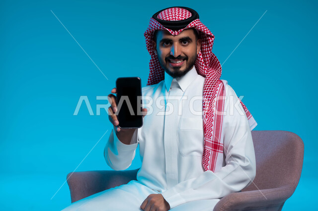 السرور والسعادة باستخدام الأجهزة والتقنيات الحديثة والمتطورة، بورتريه لرجل عربي خليجي سعودي مبتسم يرتدي الثوب والشماغ التقليدي يقوم بعرض هاتف محمول بشاشة سوداء فارغة، خلفية زرقاء