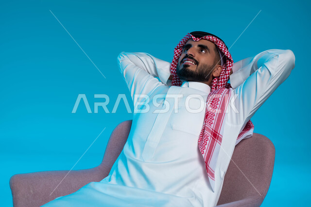 شرود الذهن و التمعن، الشعور بالتعب و الخمول والحاجة إلى النوم, بورتريه لرجل عربي خليجي سعودي يرتدي الزي التقليدي يجلس على كرسي مريح يضع يديه وراء رأسه بتعابير التأمل والتفكر، إيماءات الراحة والاسترخاء، النظر للأعلى إلى شيء ما، خلفية زرقاء.