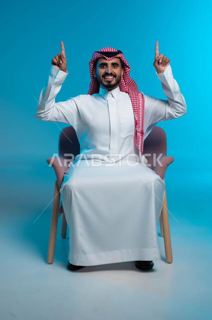 رفع اليدين إلى الأعلى والنظر إلى الكاميرا, البشاشة والسرور وتحديد شيء معين, بورتريه لشاب عربي خليجي سعودي مبتسم يرتدي الثوب والشماغ التقليدي يجلس على الكرسي ويشير بإصابع السبابة إلى شيء ما, خلفية زرقاء
