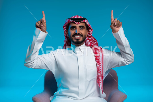 البشاشة والسرور وتحديد شيء معين, رفع اليدين إلى الأعلى والنظر إلى الكاميرا, بورتريه مقرب لشاب عربي خليجي سعودي مبتسم يرتدي الثوب والشماغ التقليدي يجلس على الكرسي ويشير بإصابع السبابة إلى شيء ما, خلفية زرقاء