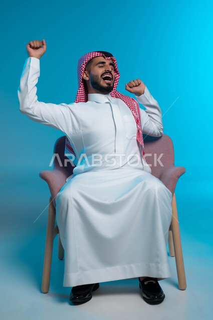 مفهوم التعب والحاجة إلى الراحة, الشعور بالنعاس والرغبه في النوم ، بورتريه لشاب عربي خليجي سعودي يرتدي الشماغ والثوب التقليدي يرفع يديه بإيماءات التثاؤب، خلفية زرقاء