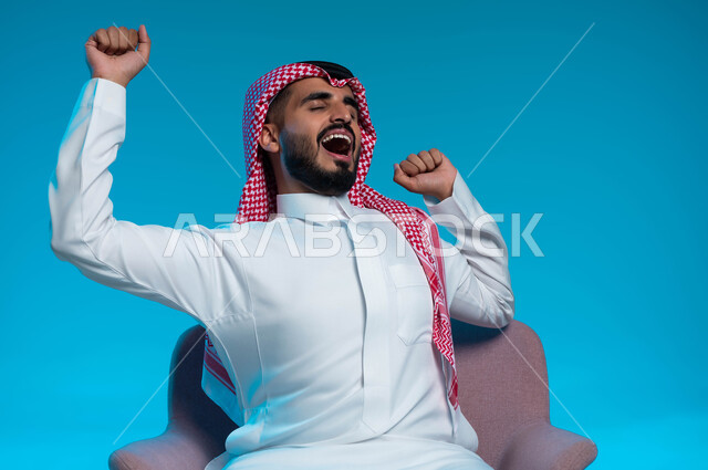 مفهوم التعب والحاجة إلى الراحة, الشعور بالنعاس والرغبه في النوم ، بورتريه لشاب عربي خليجي سعودي يرتدي الشماغ والثوب التقليدي يرفع يديه بإيماءات التثاؤب، خلفية زرقاء