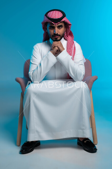 The concept of intelligence and strong will, masculinity, ambition and self-confidence, interest in elegance and external appearance, a portrait of a Saudi Gulf Arab young man wearing a shemagh and traditional dress, looking at the camera with gestures of contemplation and concentration, a blue background.