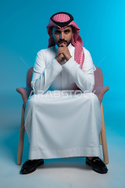 Attention to elegance and external appearance, the concept of intelligence and strong will, masculinity, ambition and self-confidence, a portrait of a Saudi Gulf Arab young man wearing a shemagh and traditional dress, looking at the camera with gestures of contemplation and concentration, a blue background.