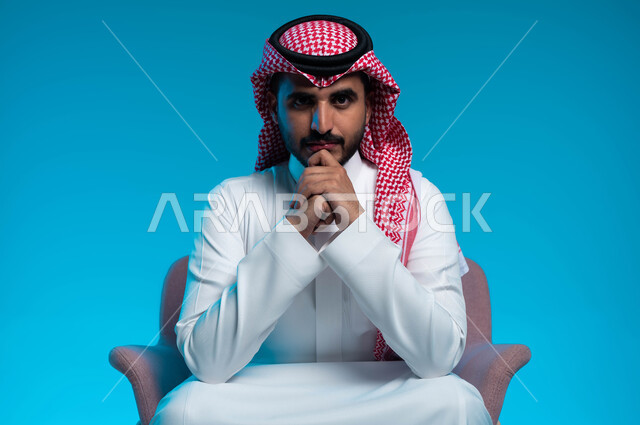 مفهوم الذكاء والإرادة القوية، الرجولة والطموح والثقة بالنفس، الاهتمام بالأناقة والمظهر الخارجي، بورتريه لشاب عربي خليجي سعودي يرتدي الشماغ والثوب التقليدي ينظر إلى الكاميرا بإيماءات التمعن والتركيز، خلفية زرقاء.