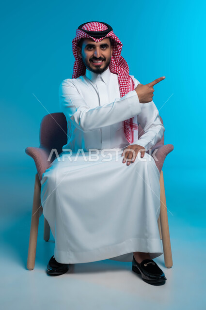 البشاشة والسرور وتحديد شيء معين, الإشارة إلى مكان ما والنظر إلى الكاميرا, بورتريه لشاب عربي خليجي سعودي مبتسم يرتدي الثوب والشماغ التقليدي يجلس على الكرسي ويشير بإصبع السبابة إلى شيء ما, خلفية زرقاء