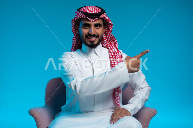الإشارة إلى مكان ما والنظر إلى الكاميرا, البشاشة والسرور وتحديد شيء معين, بورتريه لشاب عربي خليجي سعودي مبتسم يرتدي الثوب والشماغ التقليدي يجلس على الكرسي ويشير بإصبع السبابة إلى شيء ما, خلفية زرقاء