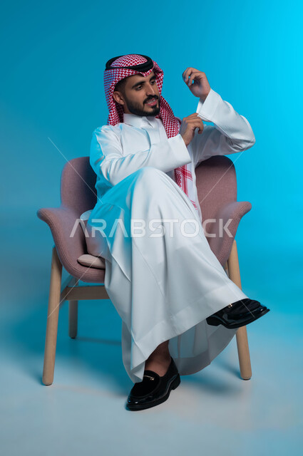 مفهوم الشياكة والأناقة والرجولة ،بورتريه لشاب عربي خليجي سعودي مبتسم يرتدي الزي التقليدي يجلس على كرسي مريح ويقوم بتعديل الثوب الابيض، ايماءات السرور و البشاشه، الاهتمام بالمظهر الخارجي، خلفية زرقاء .