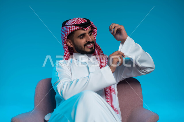 Attention to external appearance, the concept of elegance, elegance and masculinity, a portrait of a smiling Saudi Gulf Arab young man wearing traditional clothing, sitting on a comfortable chair and adjusting the white dress, gestures of happiness and cheerfulness, blue background.