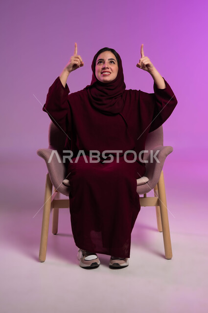 النظر الى الاعلى بإيماءات السرور، إشارات وحركات يدين تدل على هذا المكان (هنا)، تحديد موقع معين بإيماءات البشاشة، بورتريه لامرأة عربيه خليجيه سعوديه مبتسمه محجبه ترتدي العباءة الملونة تجلس على كرسي مريح وترفع اصابعها وتشير الى شيء ما، خلفية ملونة .