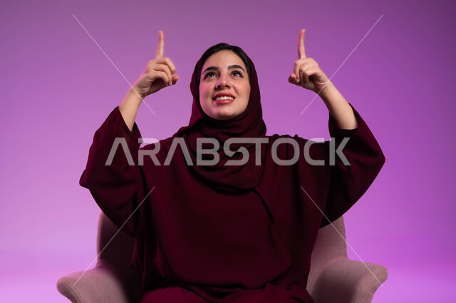 تحديد موقع معين بإيماءات البشاشة، النظر الى الاعلى بإيماءات السرور، إشارات وحركات يدين تدل على هذا المكان (هنا)، بورتريه مقرب لامرأة عربيه خليجيه سعوديه مبتسمه محجبه ترتدي العباءة الملونة تجلس على كرسي مريح وترفع اصابعها وتشير الى شيء ما، خلفية ملونة .