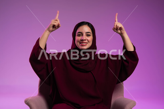 Looking at the camera with gestures of pleasure, gestures and hand movements indicating this place (here), identifying a specific location with gestures of joy, a close-up portrait of a smiling, veiled Saudi Arabian Gulf woman wearing a colored abaya, sitting on a comfortable chair, raising her fingers and pointing upwards, a colored background.