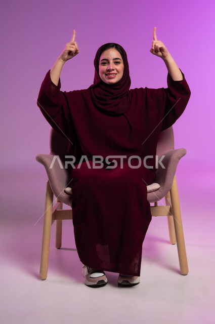 إشارات وحركات يدين تدل على هذا المكان (هنا)، تحديد موقع معين بإيماءات البشاشة، النظر الى الكاميرا بإيماءات السرور، بورتريه لامرأة عربيه خليجيه سعوديه مبتسمه محجبه ترتدي العباءة الملونة تجلس على كرسي مريح وترفع اصابعها وتشير الى الاعلى، خلفية ملونة .