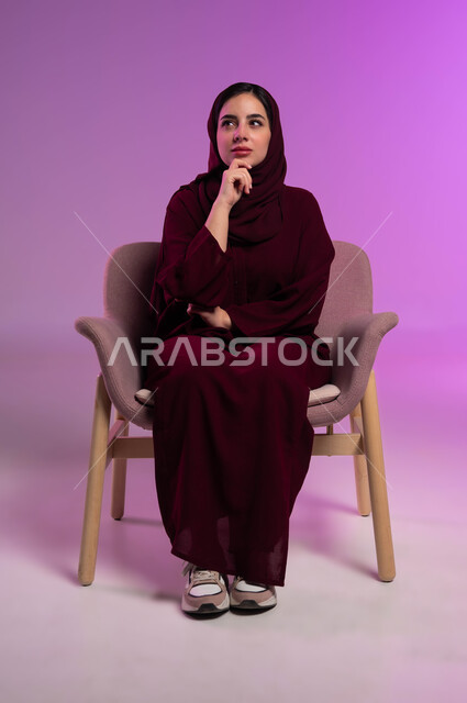 شرود الذهن والتأمل والتخطيط للمستقبل، التفكير لأيجاد حل مشكلة ما، بورتريه لامرأة عربيه خليجيه سعوديه محجبه ترتدي العباءة الملونة تجلس على كرسي مريح وتنظر الى الأعلى بإيماءات التمعن والتركيز، مواجهة المشكلات والتحديات في الحياة العملية، خلفية ملونة